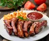 The image shows a plate of barbecued ribs accompanied by french fries and a side of ketchup garnished with a parsley leaf with fresh tomatoes and herbs in the background