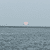A large pinkish sun hangs low over the horizon above a rippled body of water with a hint of land in the distance.