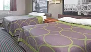 This image shows a hotel room with two twin beds covered with purple patterned bedspreads, a framed picture on the wall, and a simple interior decor.