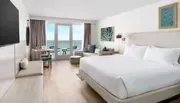 The image shows a modern hotel room with a large bed, a seating area, and a balcony overlooking the ocean.
