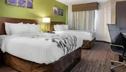 The image shows a tidy and modern hotel room with two beds featuring white linens with black floral patterns, a desk with a chair, and green and purple wall art above each bed.