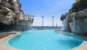 An outdoor pool with a cascading artificial waterfall, overlooking the sea, set against a clear sky.
