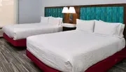 This image shows a modern hotel room with two neatly-made beds, a shared headboard with built-in nightstands, and a patterned aqua-blue backdrop.
