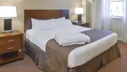 The image shows a neatly made hotel room bed with white linens and a brown bedspread, flanked by matching nightstands with lamps.
