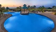 An inviting resort swimming pool, with a gazebo in the center and lounge chairs around, overlooks the beach at sunset.