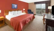 The image shows a neatly arranged hotel room with a king-sized bed, a red accent wall, contemporary furnishings, and a view outside through a glass door.