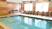 An indoor rectangular swimming pool is surrounded by brown chairs and tall windows, set within a room with tan brick walls.