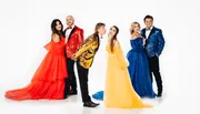 Five people wearing colorful, formal attire pose in front of a white background, with two couples on the sides and a man in the center playfully pouting towards a woman in yellow.