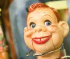 The image shows a close-up of a vintage-looking toy with a smiling freckled face and blue eyes accompanied by blurred background figures