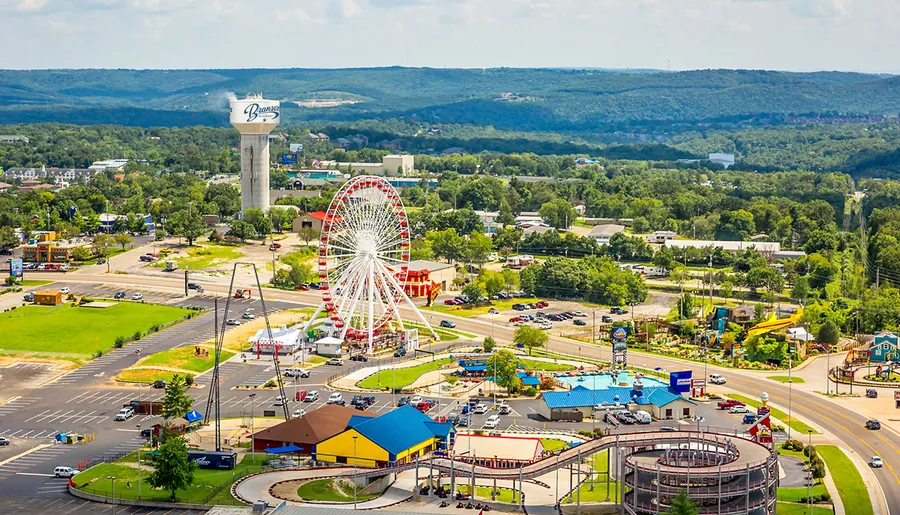 The image shows an aerial view of a vibrant amusement park, featuring a large Ferris wheel, roller coaster tracks, and colorful buildings, set against a backdrop of lush green hills.