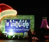 The image shows a festive entrance to The Trail of Lights with a large brightly-lit sign a decorated structure that resembles a Santa hat and a tower adorned with colorful lights in the background at night