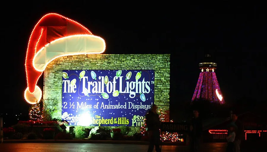 The image shows a festive entrance to 'The Trail of Lights' with a large, brightly-lit sign, a decorated structure that resembles a Santa hat, and a tower adorned with colorful lights in the background at night.