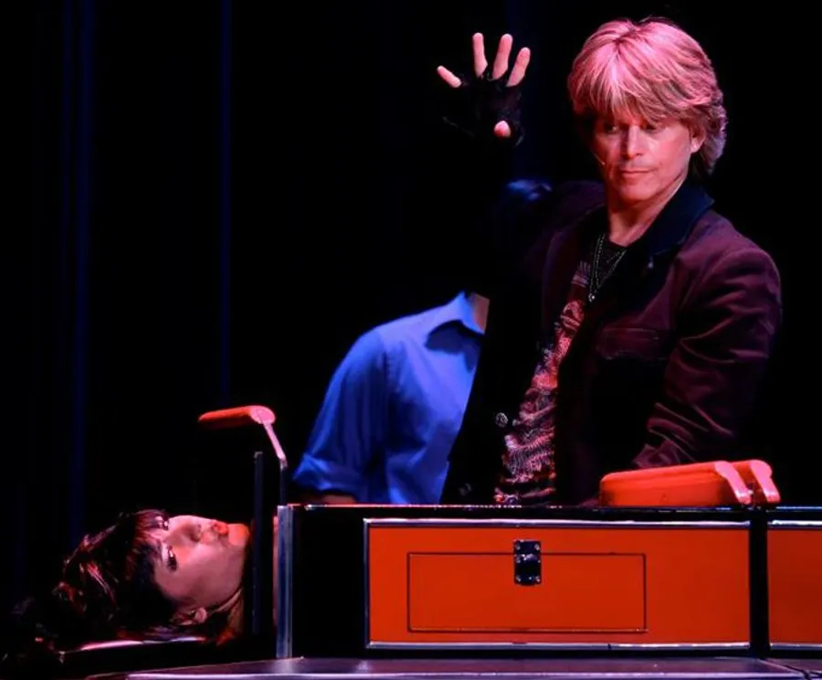 A magician is performing a classic sawing a person in half illusion on stage, with a woman lying in the box and the magician gesturing dramatically with his hand raised.