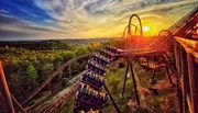 A roller coaster train ascends a track against a dramatic sunset sky, offering a picturesque view of the surrounding landscape.