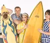 Four people are joyfully posing together with a giant yellow surfboard against a white background
