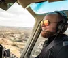 A smiling person wearing sunglasses and a headset is looking out the window of a helicopter observing a landscape from above
