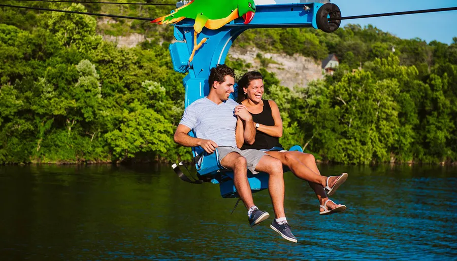 A man and a woman are smiling and enjoying a chairlift ride over a body of water, surrounded by lush greenery.