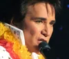 A person is performing onstage with a microphone dressed in an Elvis Presley-inspired outfit adorned with a colorful lei