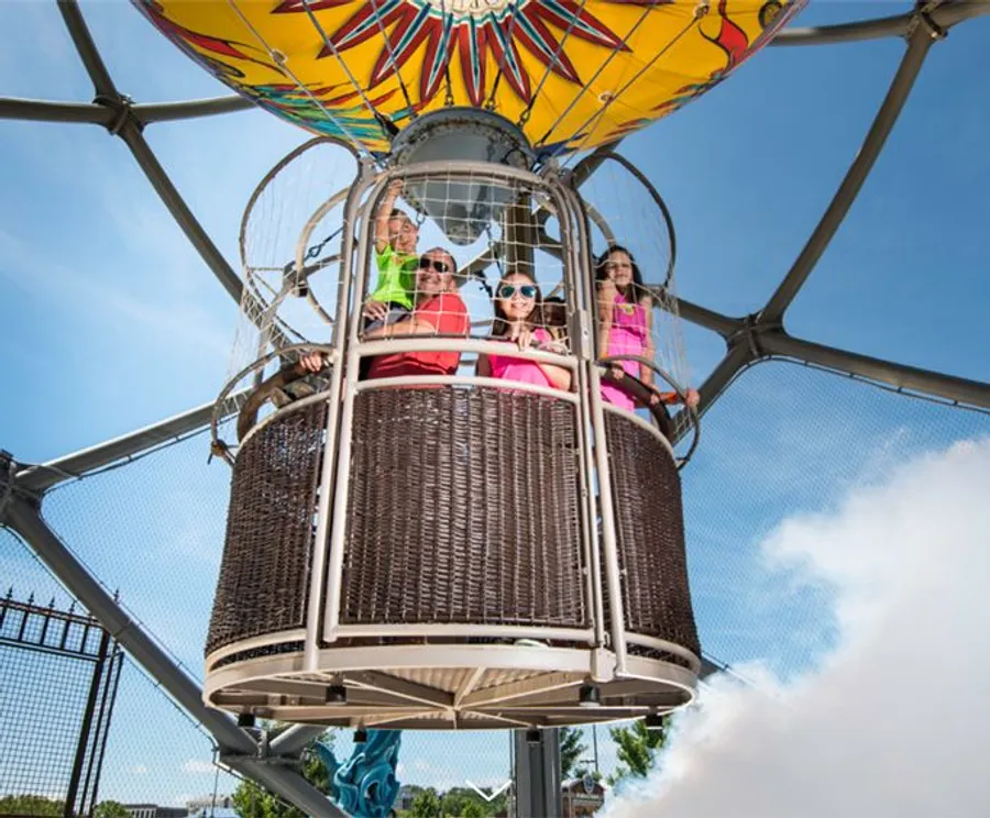 The image shows a group of people enjoying a ride in a colorful, decorative hot air balloon basket at a theme park or fairground under a clear blue sky.