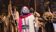 The image depicts actors in historical or biblical costumes performing in a theatrical production, with one prominently featured man in a red shawl and beard looking upwards.
