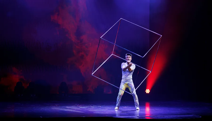 A performer on stage manipulates an intricate geometric shape under dramatic red and blue stage lighting.
