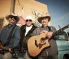 Three musicians stand confidently in front of a vintage blue pickup truck holding their guitars exuding a classic Americana vibe