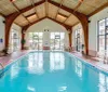 The image shows an indoor swimming pool with a vaulted ceiling large windows and several chairs along the poolside