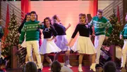 A group of people are performing on stage in festive holiday sweaters, exuding a cheerful Christmas spirit.