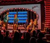 The image shows a vibrant game show set with a group of dancers performing in the foreground and the audience along with a panel of seated guests looking on