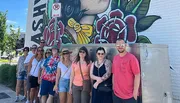 A group of people is posing for a photo in front of a large mural with vibrant artwork on a sunny day.