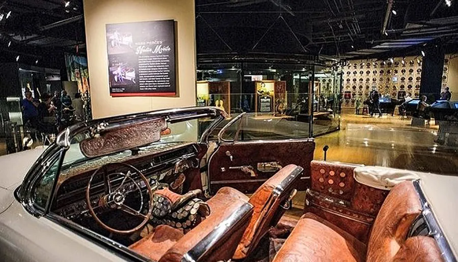 The image shows a vintage car on display inside a museum with visitors in the background looking at various exhibits.