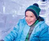 A child is joyfully sliding down a clear ice slide wearing winter clothes and a big smile
