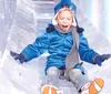 A child is joyfully sliding down a clear ice slide wearing winter clothes and a big smile