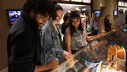 A group of people is smiling and looking at exhibits, possibly within a museum or gallery, where a guitar is displayed under glass.