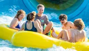 A group of friends is laughing and enjoying a sunlit ride down a water slide in a yellow raft.