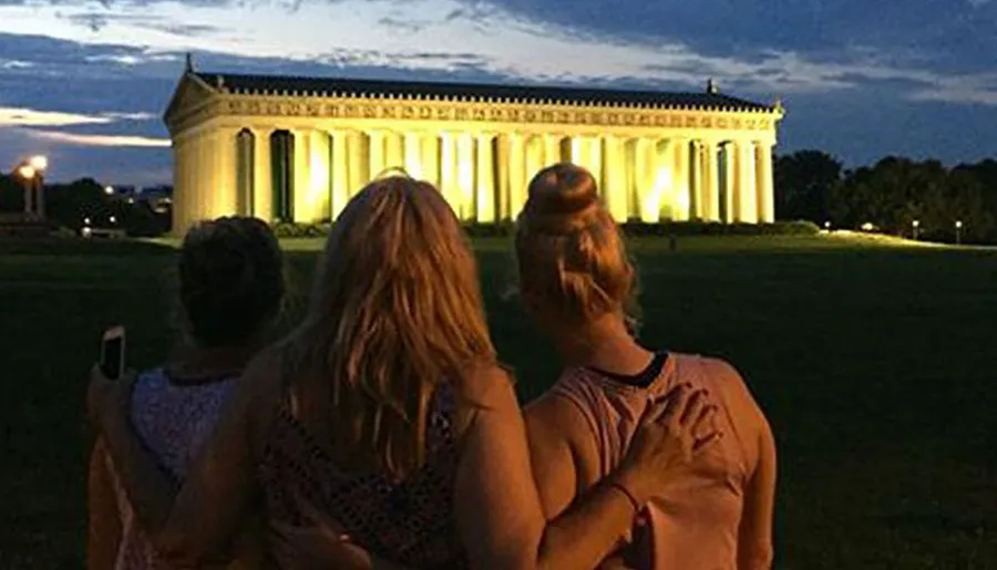 Two individuals are admiring the illuminated Parthenon replica at dusk in Nashville's Centennial Park.