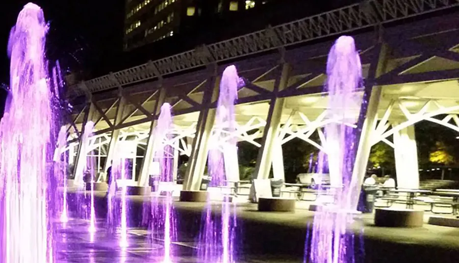 The image shows a series of water fountains illuminated by purple lights at night, with tables and chairs in the foreground and an architectural structure with white beams in the background.