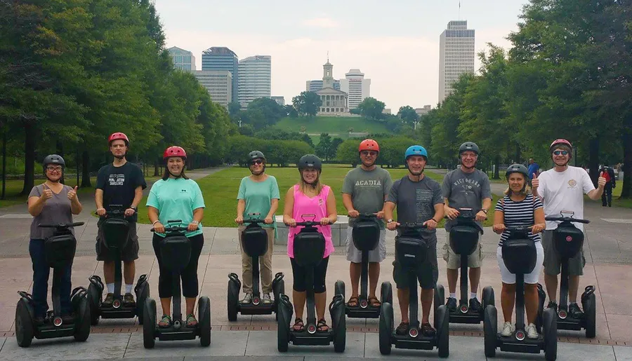 A group of people wearing helmets is posing for a photo while standing on Segways with a backdrop of greenery and city buildings.