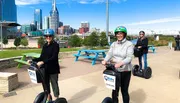Two individuals are smiling for the camera while riding Segways on a path with the Nashville skyline in the background.