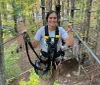 A smiling person is equipped with a harness and safety gear on a high ropes course among trees