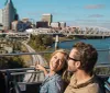 A couple is enjoying a scenic view from the top deck of a tour bus with a city skyline and a bridge in the background