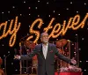 A man stands on stage with arms outstretched in front of a lit sign that displays the name Ray Stevens with musicians in the background