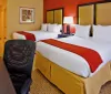 The image displays a bright and neatly arranged hotel room with two queen-sized beds a work desk and chair and warm color accents