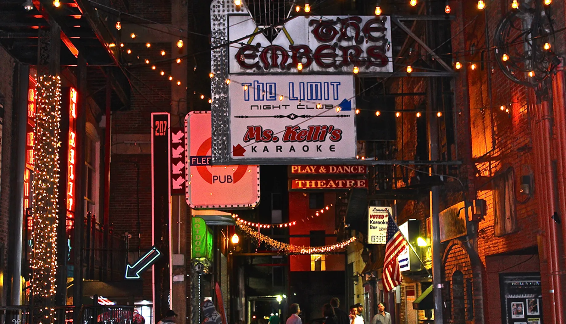 The image shows a vibrant city alley lined with various neon signs for bars, nightclubs, and a karaoke place, creating a lively nightlife ambiance.