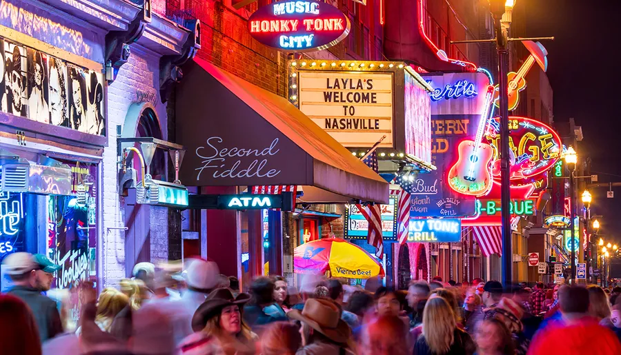 The image captures a lively and crowded street at night, illuminated by the colorful neon signs of bars and music venues, synonymous with the vibrant nightlife of Nashville, Tennessee.
