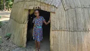 A smiling child is standing in the doorway of a thatch-roofed hut.