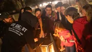 A group of people gather around a lantern at night, listening attentively to a person gesturing dramatically, likely on a ghost tour.