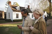 A smiling couple is looking at their smartphones while walking together on a sidewalk in front of a white historic-looking house.