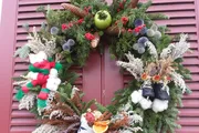 The image shows a festive Christmas wreath adorned with greenery, colorful decorations including red berries and pinecones, and hints of winter themes such as cotton resembling snow, hanging on a reddish-brown door.