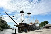 A replica of a historical sailing ship is docked at a wooden pier under a sky streaked with wispy clouds.